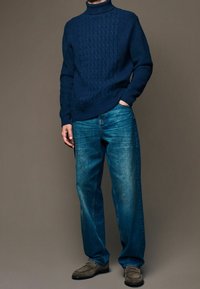 Homme portant un pull col roulé en tricot torsadé bleu marine, un jean bleu à coupe décontractée et des mocassins en suède gris, se tenant avec une main dans la poche.