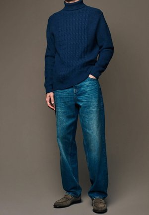 Mann in navyblauem, grob gestricktem Rollkragensweicher, lässigen blauen Jeans und grauen Wildleder-Pantoletten, steht mit einer Hand in der Tasche.