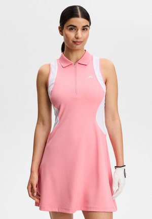 Frau trägt ärmelloses Golfkleid in Pink und Weiß mit Reißverschlusskragen und weißem Golfhandschuh an der rechten Hand vor einfarbigem Hintergrund.