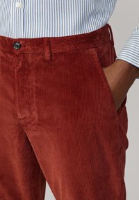 Pantalones de pana marrones con acanaladuras verticales, que presentan una cinturilla con cierre de botón y bolsillos laterales, combinados con una camisa a rayas azul.