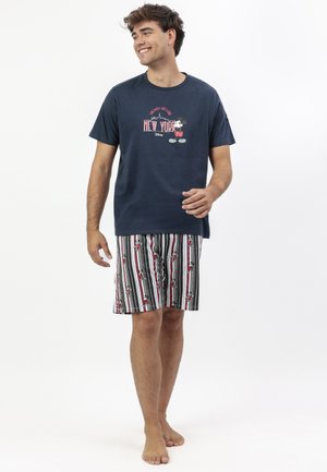 Hombre sonriente vistiendo una camiseta azul marino de Mickey Mouse con el texto New York y pantalones cortos a rayas con estampados de Mickey Mouse, caminando descalzo sobre un fondo blanco.