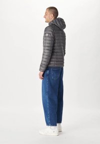 Grå pufferjacka med huva, quiltad design och logo på axeln, kombinerad med lösa blå jeans och vita sneakers.