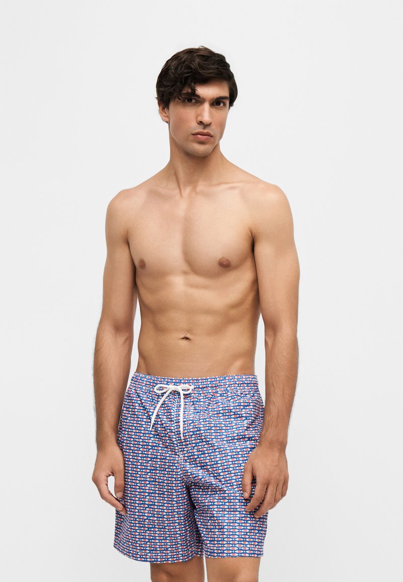 Homme torse nu aux cheveux foncés portant un short de bain bleu et rouge à motifs avec un cordon blanc, debout devant un fond clair uni.