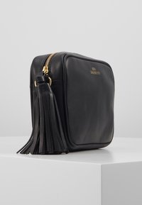 Czarna skórzana prostokątna torba crossbody z złotym zamkiem błyskawicznym, dużym skórzanym frędzlem i małym złotym logo z przodu, umieszczona na białej powierzchni.