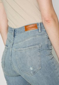 Los pantalones de mezclilla de color azul claro presentan un diseño de cintura alta con un parche de cuero marrón, mostrando un desgaste mínimo y bolsillos traseros.