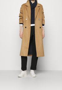 Camelivärinen villa takki, jossa on viistokaulus, kaksirivinen muotoilu ja suuret napit, kerrostettuna tummansinisen neuleen ja mustien housujen päälle.