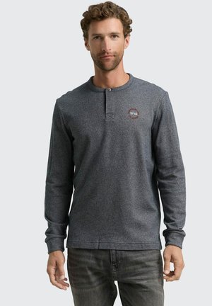 Langärmliges Henley-Shirt aus dunkelgrauem strukturiertem Stoff mit einem knopfbaren Kragen und kleinem Logodetail auf der Brust. Kombiniert mit grauen Jeans.