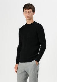 Pull en tricot noir avec un col rond, des manches longues et une coupe ajustée, associé à un pantalon gris clair à rayures.