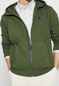 Grön zip-up hoodie i bomullsblandning, med dragsko på huvan, två sidofickor och en liten logo på bröstet. Lätt inre textur.