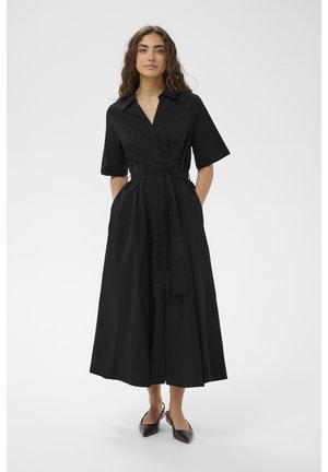 Femme portant une robe chemise midi noire à manches courtes, avec ceinture à la taille et des chaussures slingback à bout pointu, debout les mains dans les poches.