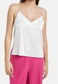 Camisole en satin blanc avec de fines bretelles, décolleté en V et détails de boutons décoratifs sur le devant. Des fentes latérales accentuent la coupe ample.