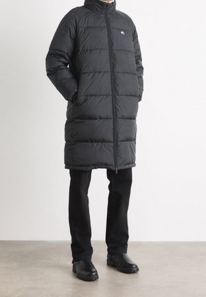 Person trägt einen langen schwarzen gesteppten Daunenmantel mit Reißverschluss, Hände in den Taschen, schwarze Hose und schwarze klobige Stiefel vor neutralem Hintergrund.