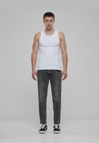 Top sin mangas blanco, jeans grises y zapatillas negras. El modelo está de pie sobre un fondo neutro, mostrando un atuendo casual con un diseño ajustado.