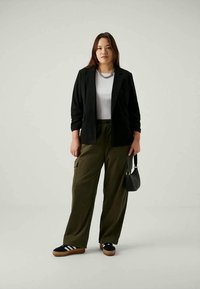 Blazer preto sobre uma t-shirt branca, calças cargo verde-azeitona, mala preta e sapatilhas pretas com riscas brancas. Fundo neutro.