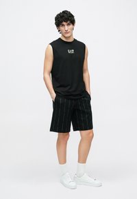 Νέος άνδρας με σγουρά μαλλιά φοράει μαύρο αμάνικο μπλουζάκι, μαύρο σορτς με σχέδια, άσπρες κάλτσες και άσπρα αθλητικά παπούτσια, στέκεται με τα χέρια στις τσέπες.