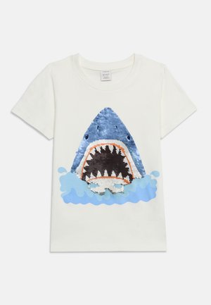 T-shirt blanc à manches courtes avec un grand motif tête de requin bleue en sequins émergeant d'une éclaboussure d'eau bleue à l'avant.