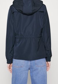 Mörkblå lättviktsjacka med huva, elastiska muddar och dragsko i midjan; slät yta, minimalistisk design, ryggvy visas.