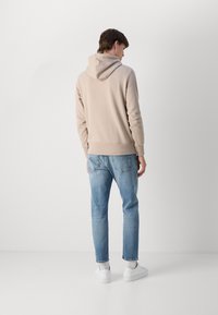Felpa beige con cappuccio e orlo a costine, abbinata a jeans azzurri. Sneakers bianche completano l'outfit, vista da dietro.
