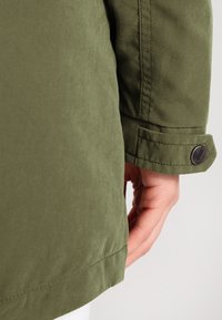 Manche de veste verte avec poignet boutonné reposant près de la main d'une personne sur un fond clair uni.