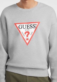 Grå crewneck genser laget av stoff, med en rød trekantet logo som har teksten "GUESS U.S.A" og "WASHED JEANS" i svart.