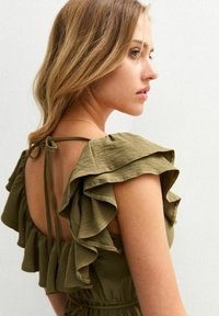 Vestido verde oliva con acentos de hombros con volantes y atado en la parte posterior. Fabricado con un tejido ligero de acabado texturizado, que presenta una cintura entallada.