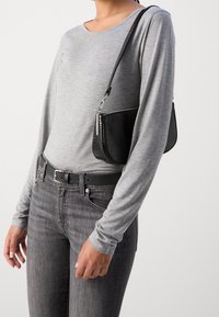 Graues Langarmoberteil, schwarzer Gürtel und dunkelgraue Jeans; hält eine schwarze Handtasche mit Krokodil-Prägung und silbernen Beschlägen.