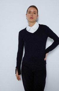 Haut en tricot à manches longues bleu marine avec un col en V et une chemise à col blanc en dessous. Présente des fentes latérales aux poignets et une texture lisse.