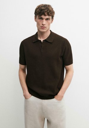 Giovane uomo con capelli ricci castani che indossa una polo a maniche corte di maglia marrone scuro e pantaloni beige chiaro, in piedi con le mani nelle tasche.