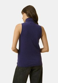 Paarse, mouwloze coltrui met een zachte textuur. Heeft een geribbelde kraag en een aansluitend ontwerp, gecombineerd met zwarte jeans.