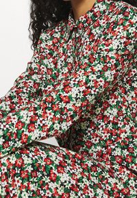 Donna che indossa una camicia a maniche lunghe e pantaloni con un denso motivo floreale rosso, bianco e verde su uno sfondo nero.