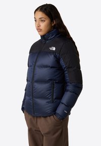 Kvinna med en marinblå och svart pufferjacka med dragkedjefickor och bruna byxor, stående med händerna i fickorna och tittande åt vänster.