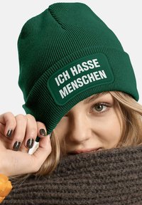 Green knit beanie featuring a rectangular patch with white text "ICH HASSE MENSCHEN." Material is textured with vertical ribs.
