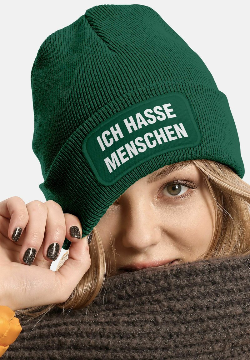 Green knit beanie featuring a rectangular patch with white text "ICH HASSE MENSCHEN." Material is textured with vertical ribs.