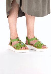 Zielone sandały z wiązaniami, teksturowanym materiałem górnym oraz białą, rowkowaną podeszwą. Kwiatowe ozdoby dodają koloru i detali.