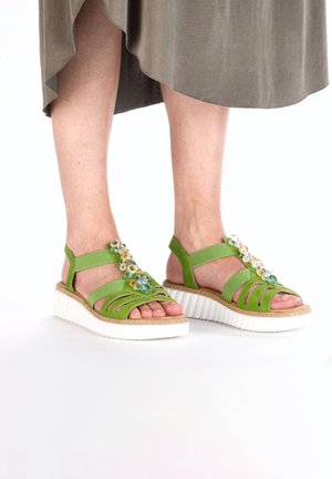 Green sandals with strappy design, textured upper material, and a white, ridged sole. Floral embellishments add color and detail.