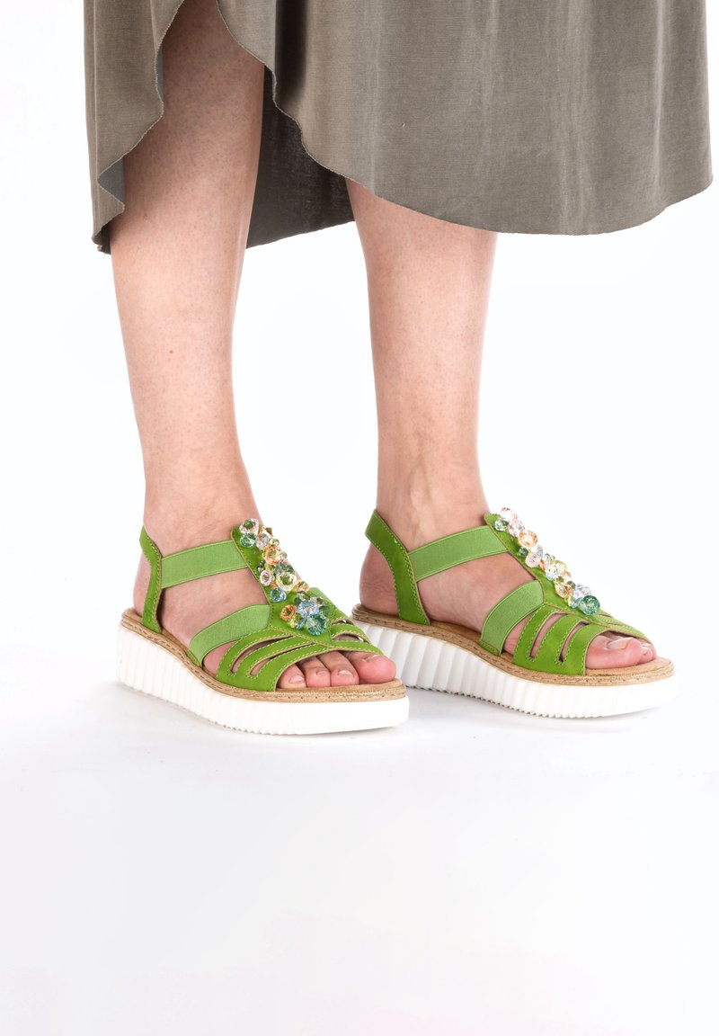 Zielone sandały z wiązaniami, teksturowanym materiałem górnym oraz białą, rowkowaną podeszwą. Kwiatowe ozdoby dodają koloru i detali.