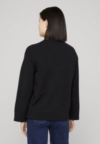 Pull noir en maille, avec un col rond, des épaules tombantes et des poignets côtelés, porté avec un jean bleu foncé.
