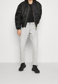 Persona vestida con una chaqueta bomber negra, camisa negra, jeans grises claros y zapatillas negras, de pie contra un fondo blanco liso.