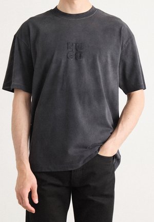 Mørkegrå bomuldst-shirt med rund halsudskæring, korte ærmer og et præget "HUGO"-logo på brystet, båret med sorte bukser.