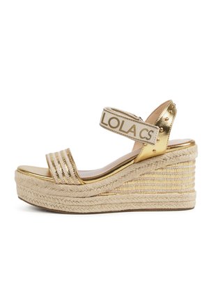 Sandali wedge dorati con piattaforma in juta intrecciata, cinturino metallico e fascia in tessuto alla caviglia con il testo "LOLA". Finitura liscia con dettagli in borchie.