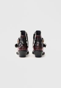 Une paire de bottines noires brillantes à talons bas, avec des boucles latérales et des zips à l'arrière, présentées de dos sur un fond blanc uni.