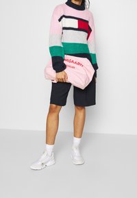 Multifarvet strikket sweater med høj krave, der har pink, grønne og marineblå striber. Holder en pink taske med røde bogstaver. Sorte shorts, hvide sneakers.