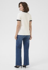 T-shirt blanc à manches courtes avec des bordures noires, associé à un jean bleu à jambes larges. Tissu lisse avec une coupe décontractée et des détails de couture visibles.