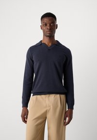 Navy knitted sweater with a V-neck and collar, featuring long sleeves and a fitted hem. Worn with beige trousers.