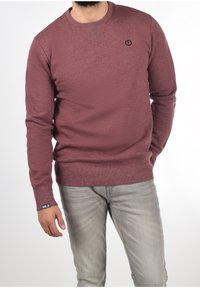 Maroon crew neck sweatshirt with textured fabric, ribbed cuffs, and a small logo on the chest. Paired with light gray denim jeans.