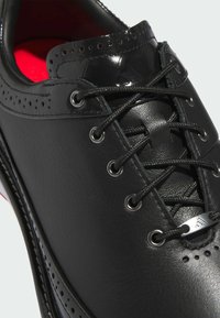 Czarny skórzany but o gładkim wykończeniu, z detalami typu brogue, błyszczącymi przelotkami, czarnymi sznurówkami oraz kontrastowym czerwonym wnętrzem.