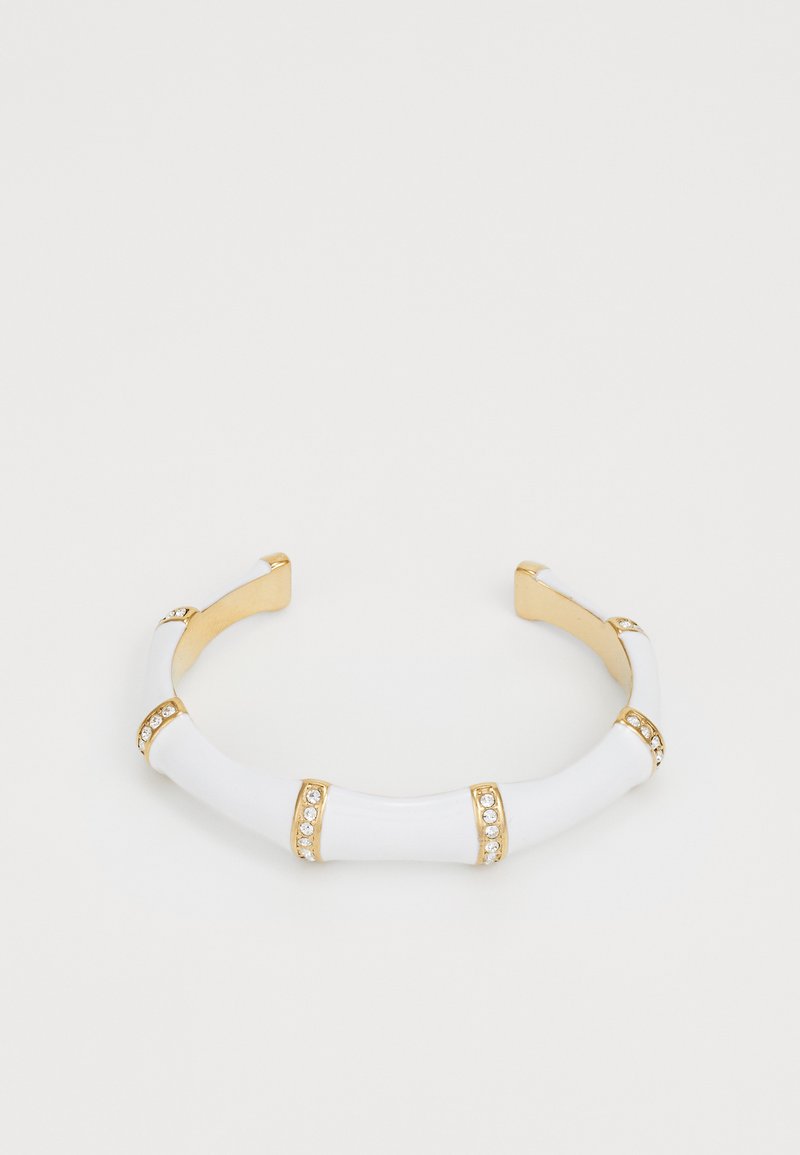 Zelta un baltas bangles rotas ar izliektām sekcijām, kas rotātas ar mazām kristālu akcentiem un gludu keramikas līdzīgu tekstūru.