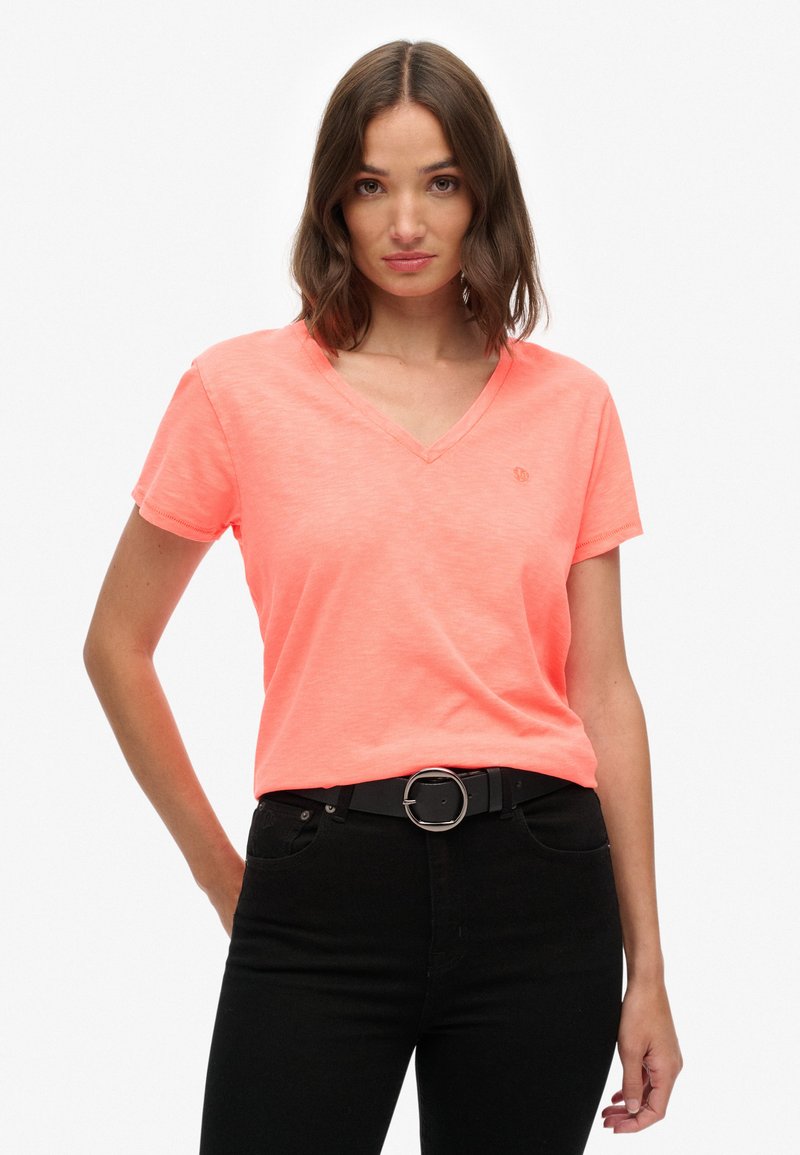 V-hals, kortemouwen t-shirt van helder neonroze stof, met een subtiele logo-detail, gedragen met een zwarte riem en donkere jeans.