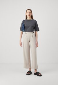 T-shirt en coton gris, pantalon ample crème avec des accents de boutons, et sandales noires. Le modèle porte un sac à bandoulière texturé bleu.