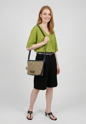 Hymyilevä nainen vihreässä topissa ja mustissa polvipituisissa shortseissa kantaa kuvioitua olkalaukkua ja pitää mustia korkokenkäisiä sandaaleja harmaalla taustalla.